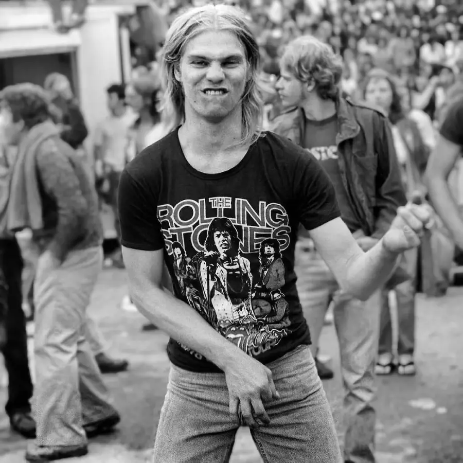 A young man with long hair wearing a Rolling Stones t-shirt and jeans pretends to play air guitar, making a playful face at an outdoor concert, with a crowd of people in the background.
