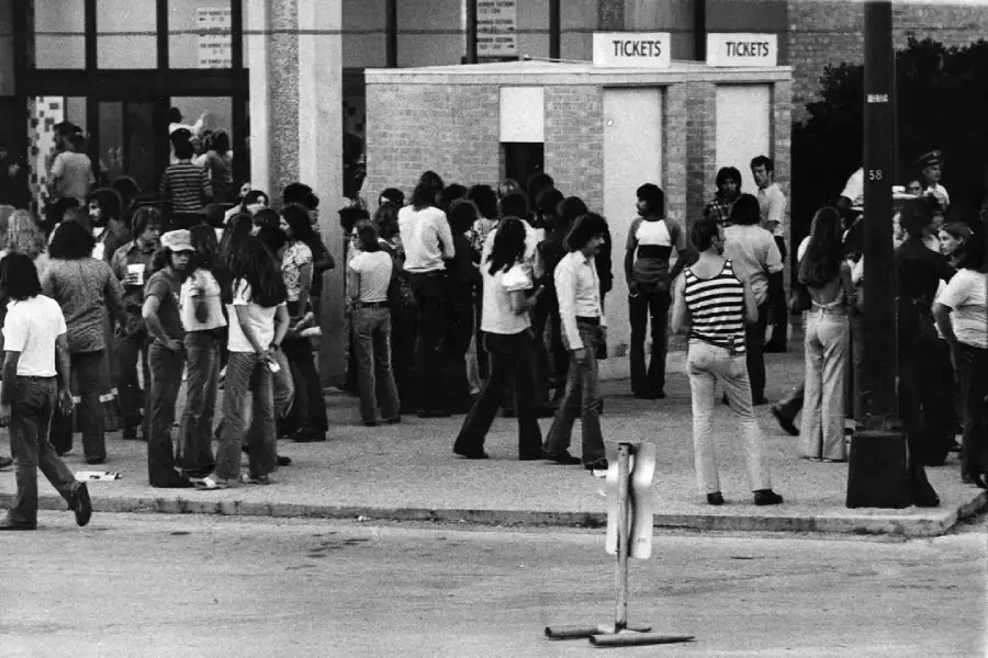 A large group of people, mostly young adults, stand and walk near a ticket booth outside a building. Many are dressed in 1970s-style clothing, and the scene appears busy and lively.