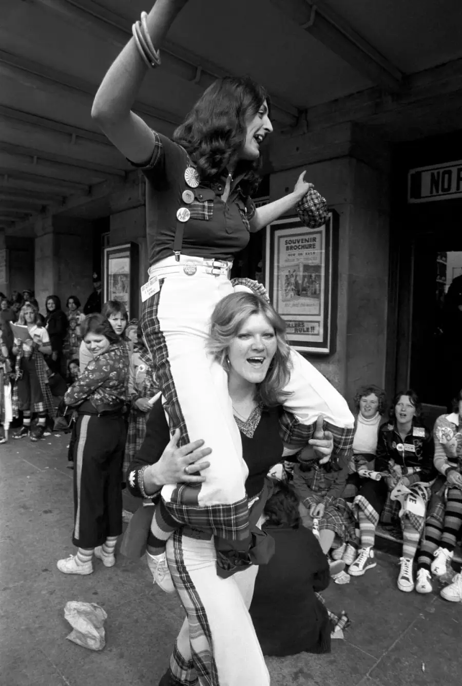 Two young women dressed in plaid clothing celebrate, with one sitting on the other's shoulders, smiling and cheering, surrounded by a crowd of similarly dressed people outside a building.