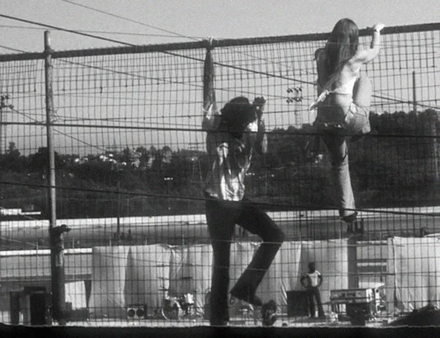 Two people climb a tall wire fence, while another person appears to watch from below. The scene is outdoors, with buildings and trees in the background and equipment visible on the ground. The image is in black and white.