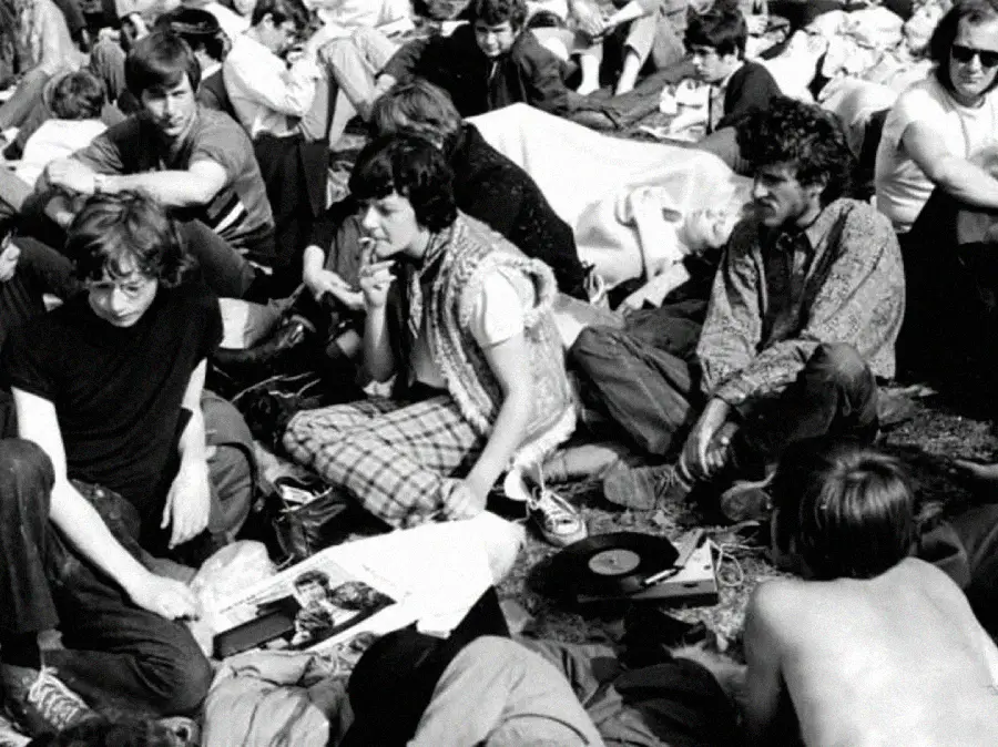 A group of young people sit closely together on the ground outdoors among blankets and records, some talking and relaxing, in a crowded, casual setting, likely at a music festival in the 1960s or 1970s.