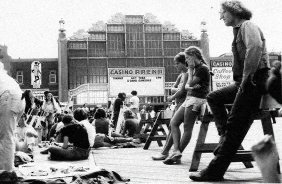 Black-and-white photo of people relaxing and walking on a crowded boardwalk in front of a large building with signs for a casino, coffee shop, and a performance by David Sandborn. Some people are seated, others are standing.