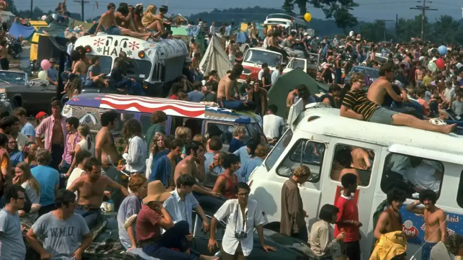 A large crowd of people at an outdoor festival gather around vehicles, many sitting on vans and buses, with tents, colorful decorations, and balloons visible in the busy, festive scene.