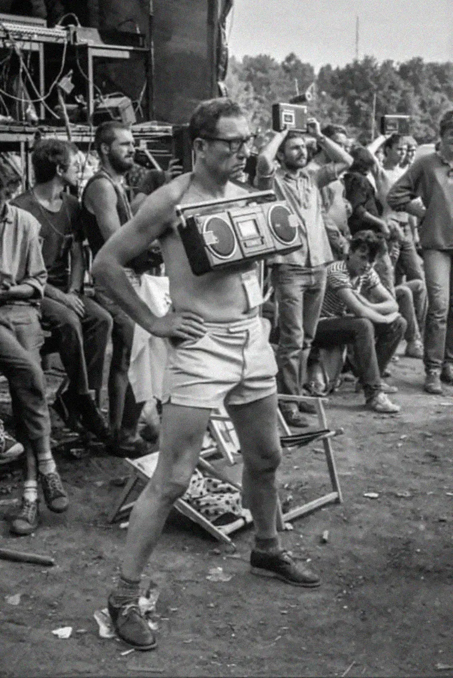 A man in shorts and glasses stands confidently with a boombox strapped to his chest at an outdoor event. People are sitting and standing around him, some holding cameras. The scene appears to be from the past.