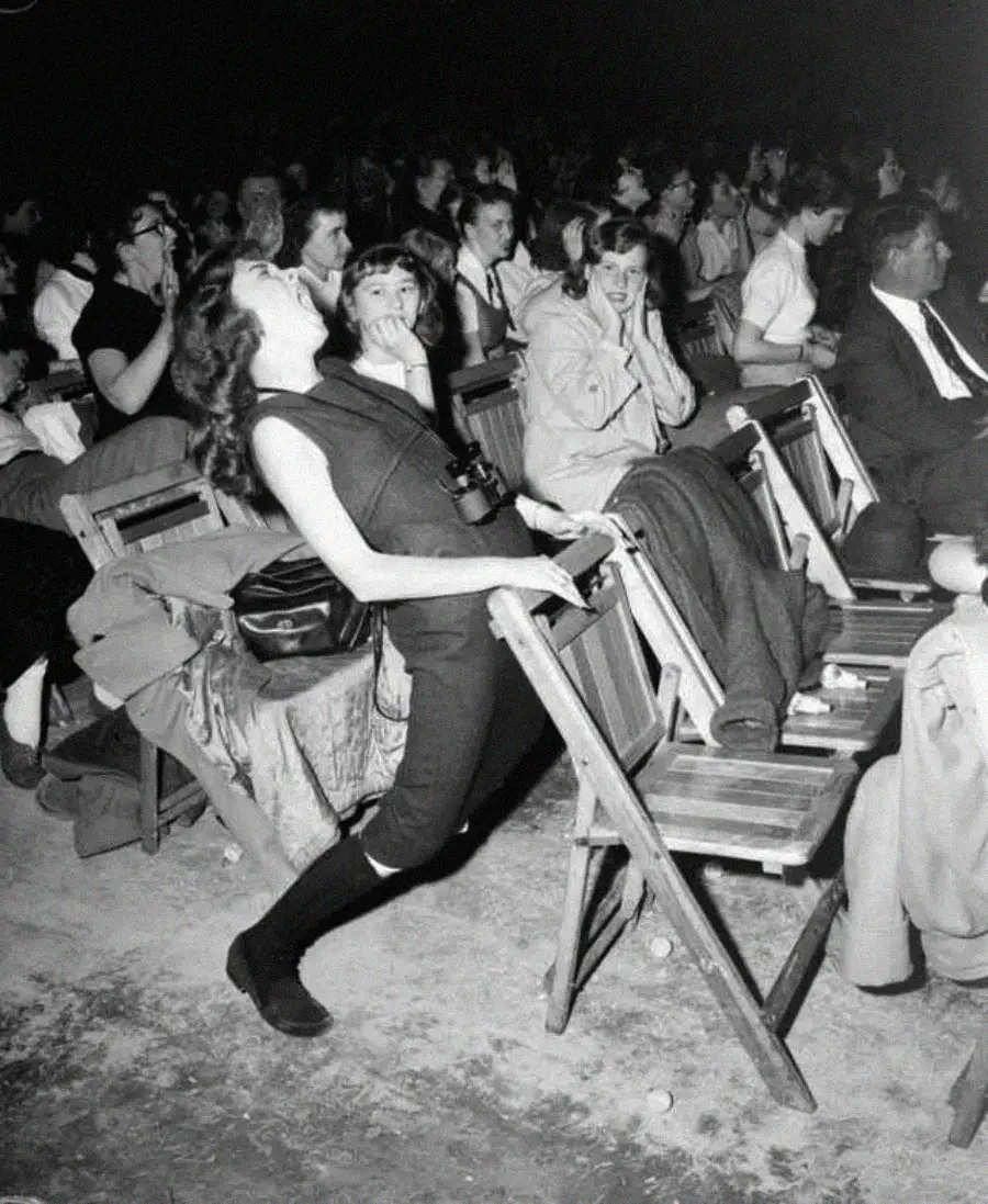 A woman leans back dramatically over a folding chair, appearing excited or overwhelmed, while others sit and watch, some with amused or surprised expressions, at a crowded event. The image is in black and white.