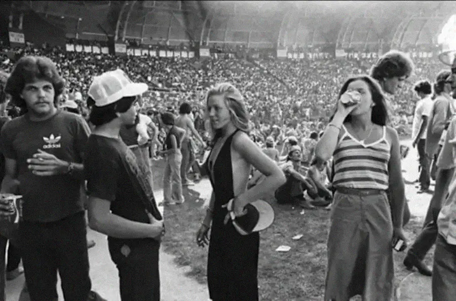 A black-and-white photo of young people standing and talking at an outdoor concert or festival, with a large crowd seated on the grass in the background. Some are holding drinks and wearing casual 1970s-style clothing.