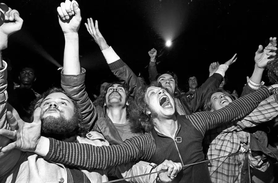 A black-and-white photo of an excited crowd at a concert, with people raising their arms, cheering, and showing expressions of joy and enthusiasm.