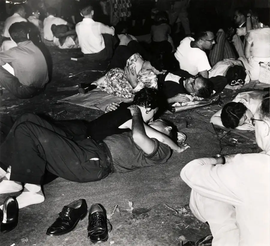 A black-and-white photo shows people lying or sitting closely together on the ground at night. A couple in the foreground embraces, while others rest or talk around them. Shoes and personal items are scattered nearby.