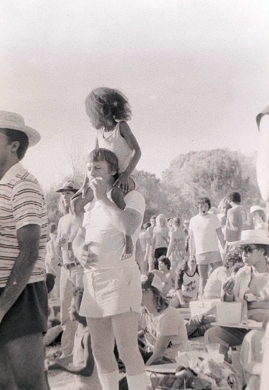 A black-and-white photo of a crowded outdoor gathering. A man in light clothing carries a young girl on his shoulders. People are sitting and standing, some wearing hats, enjoying a sunny day. Trees are visible in the background.
