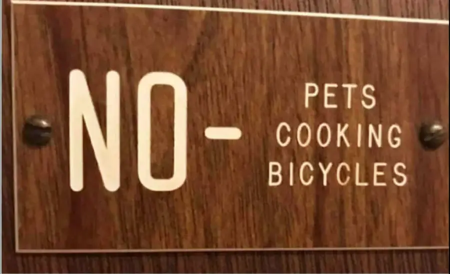 A wooden sign reads: "NO - PETS COOKING BICYCLES," with the words listed vertically, creating a humorous and confusing message.