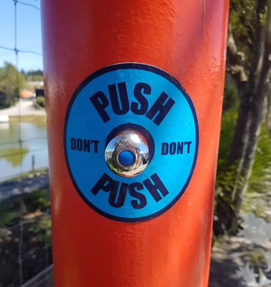 A shiny button on an orange pole with a blue label that says "PUSH" at the top and bottom, and "DON'T" on the left and right, creating conflicting instructions. Outdoor setting in the background.