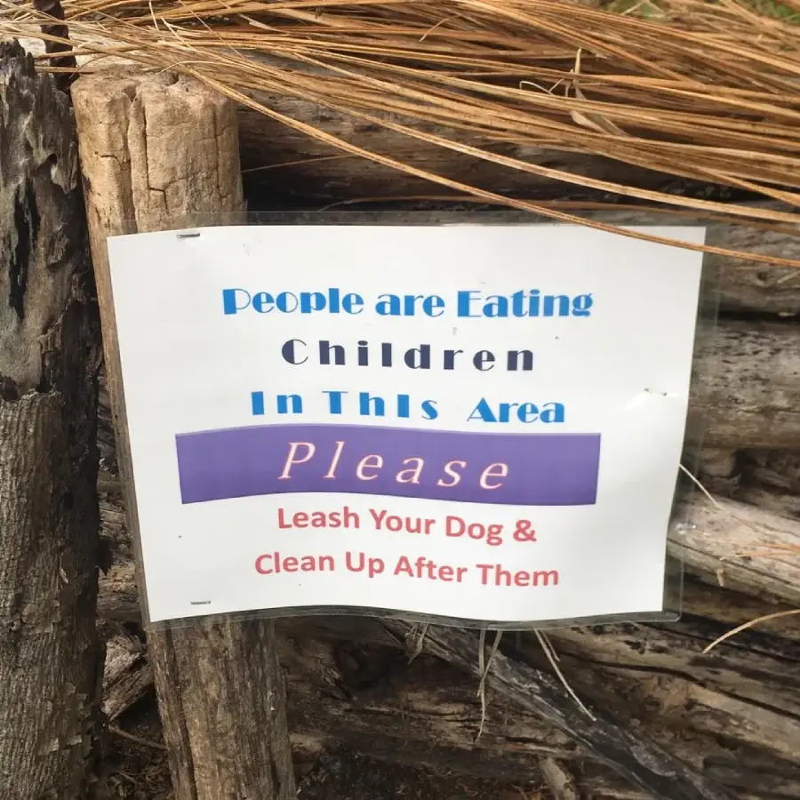 A sign humorously reads: “People are eating children in this area. Please leash your dog & clean up after them.” The odd wording creates a funny misunderstanding. The sign is posted on a wooden fence.