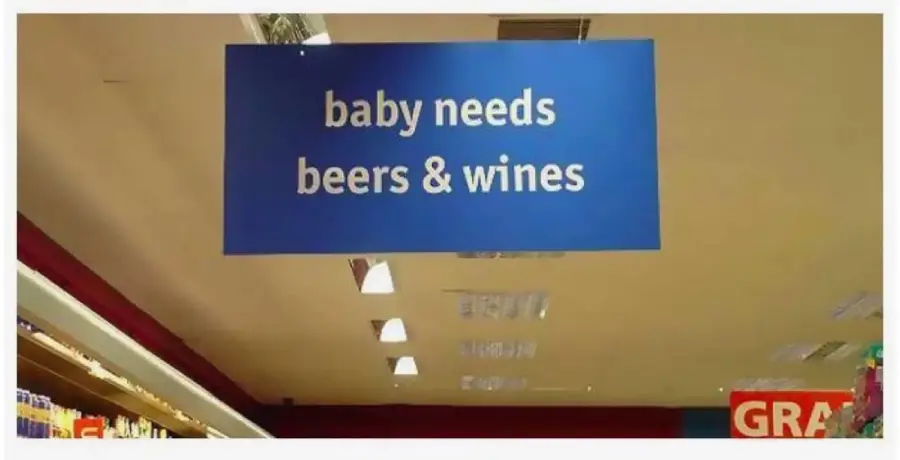 A blue store sign hanging from the ceiling reads "baby needs beers & wines," creating a humorous and confusing message due to the inappropriate pairing of baby products with alcohol.