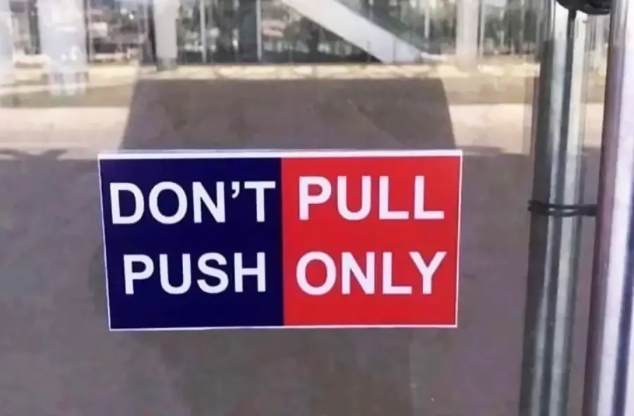 A sign on a glass door reads "DON'T PULL" on a blue background and "PUSH ONLY" on a red background, instructing people to only push the door and not pull it.
