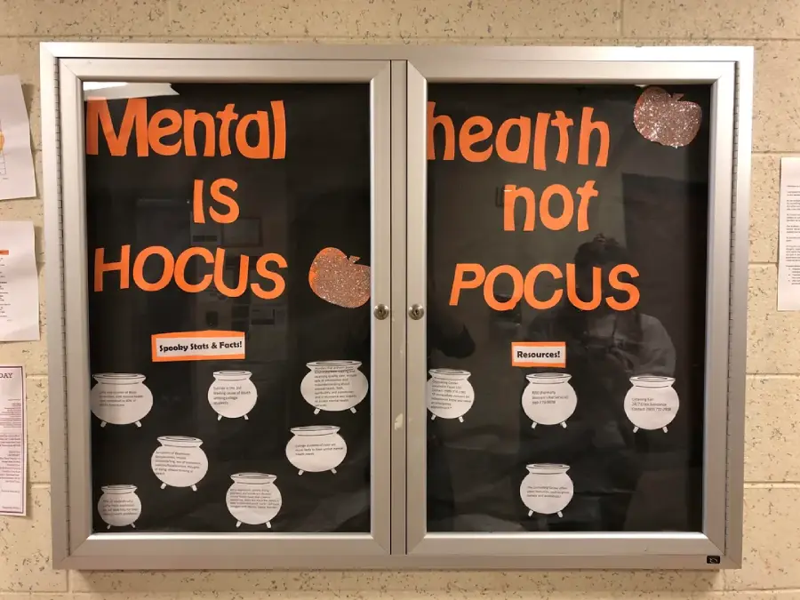 A bulletin board with the words "Mental health is not hocus pocus" in orange letters, decorated with glittery pumpkins and white cauldrons containing text on a black background.
