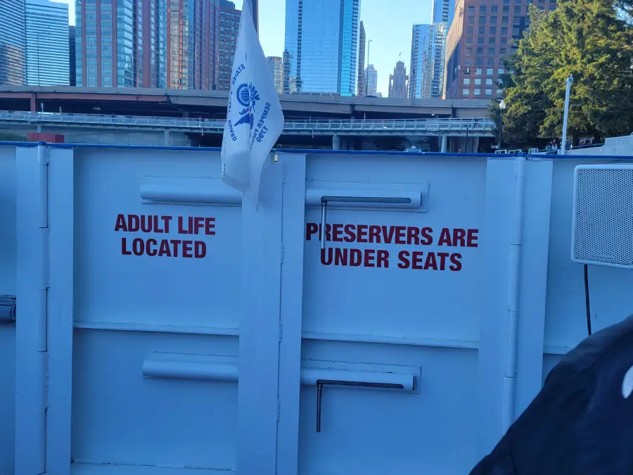 White wall on a boat with red text reading "ADULT LIFE PRESERVERS ARE LOCATED UNDER SEATS." City buildings and a flag are visible in the background.