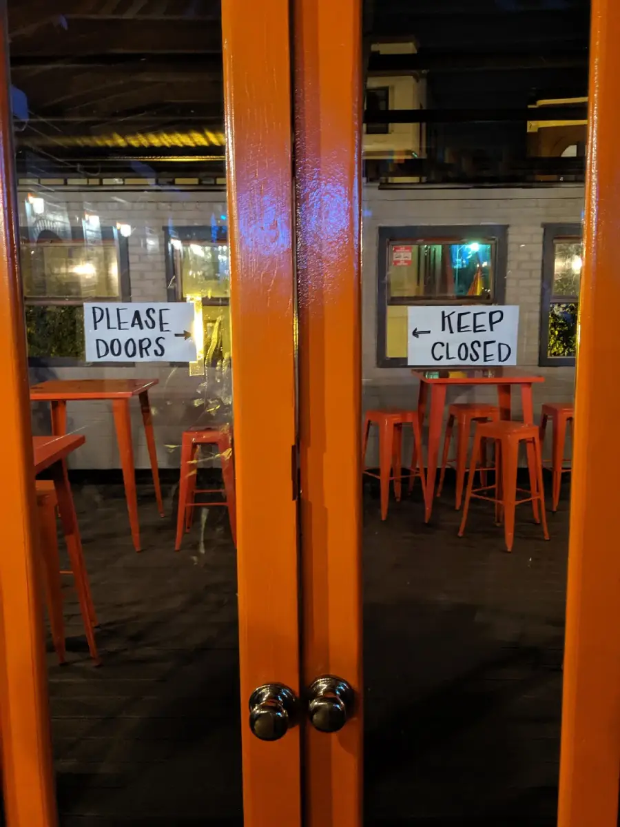 Two orange double doors with signs: "PLEASE DOORS" on the left door and "KEEP CLOSED" on the right door. Inside, orange stools and tables are visible in a dimly lit room.