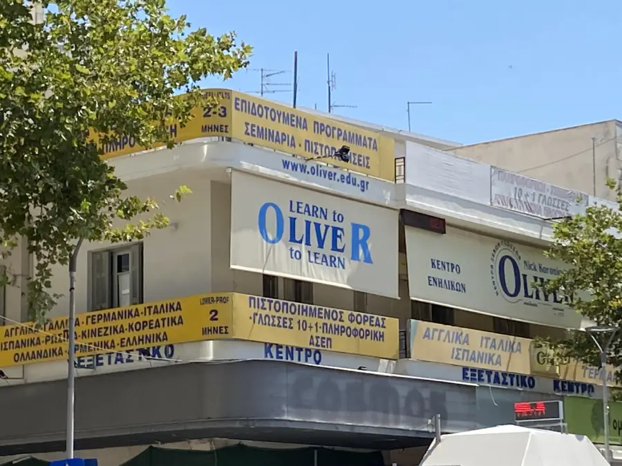 A building with multiple yellow banners advertising language courses in Greek and English, featuring a large sign that reads "LEARN TO OLIVE R TO LEARN" on the corner balcony. Trees partially shade the facade.