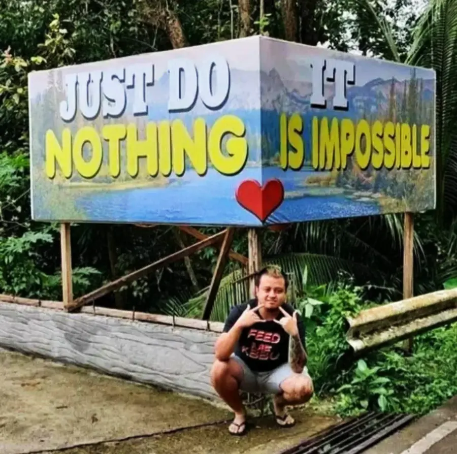 A person squats in front of a sign that reads, “JUST DO NOTHING IS IMPOSSIBLE” with some words in different sizes and colors, creating a confusing message. There are trees and greenery in the background.