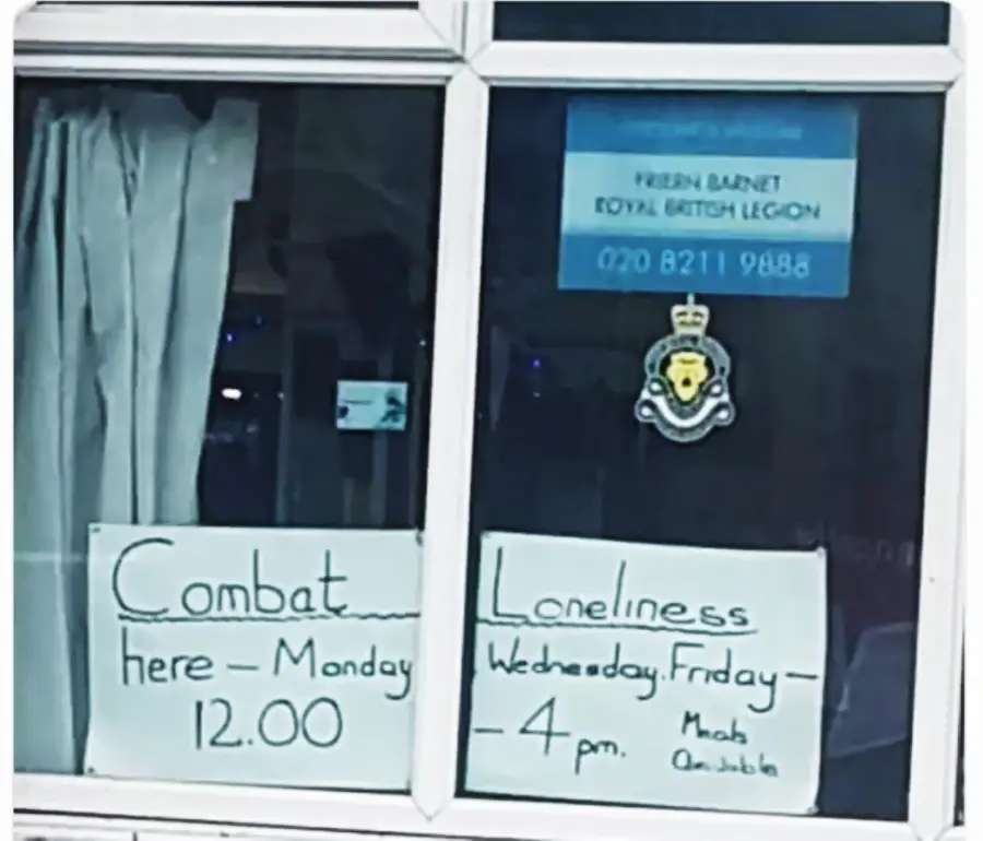 A window displays two handwritten signs: “Combat here – Monday 12.00” and “Loneliness Wednesday, Friday – 4 pm.” Above, there’s a blue sign for the Royal British Legion and a crest sticker on the glass.