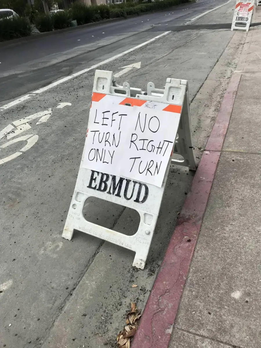 A sidewalk sign reads, “LEFT ONLY TURN, NO RIGHT TURN,” with handwritten words on white paper taped to a construction barricade labeled “EBMUD” near a street and bike lane.