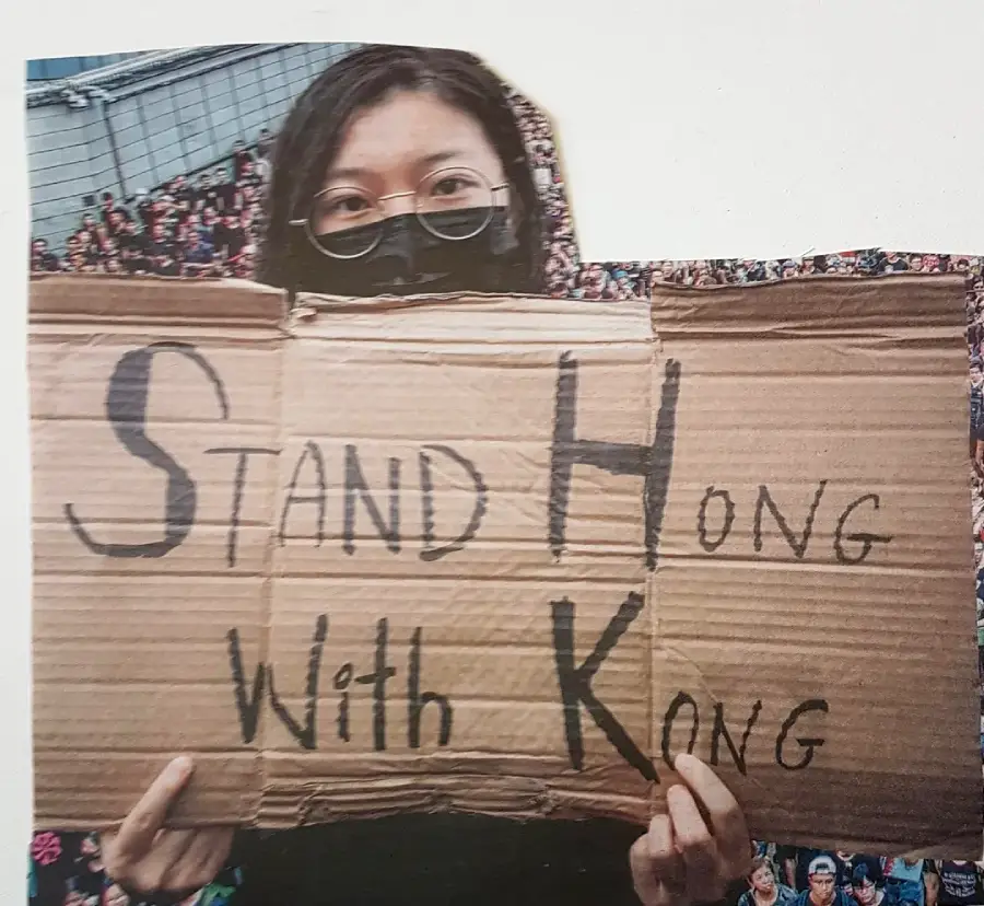 A person wearing glasses and a black face mask holds a cardboard sign reading "STAND HONG WITH KONG" in bold letters. A large crowd is visible in the blurred background.