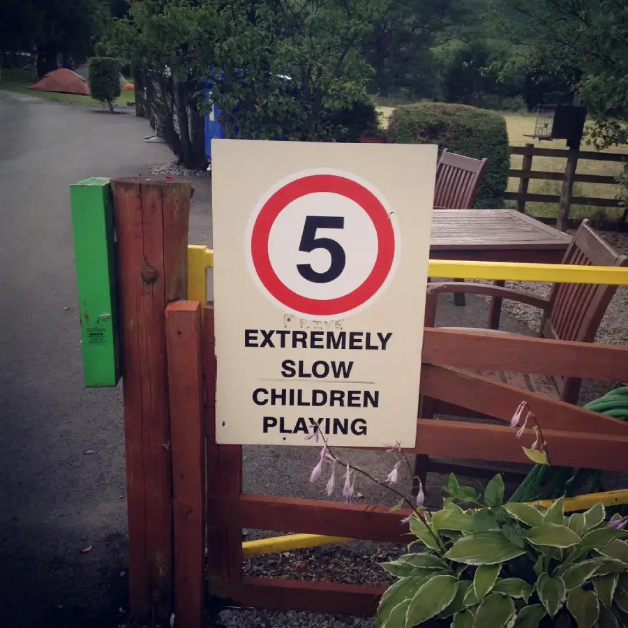 A sign next to a fence reads "5" in a red circle, with "EXTREMELY SLOW CHILDREN PLAYING" below, unintentionally suggesting the children are very slow instead of warning drivers to slow down.