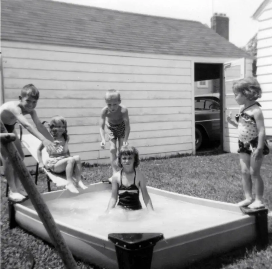 Four children in swimsuits play around a small backyard wading pool, with one child sitting in the water. A garage, a car, and a house are visible in the background. The image is black and white.