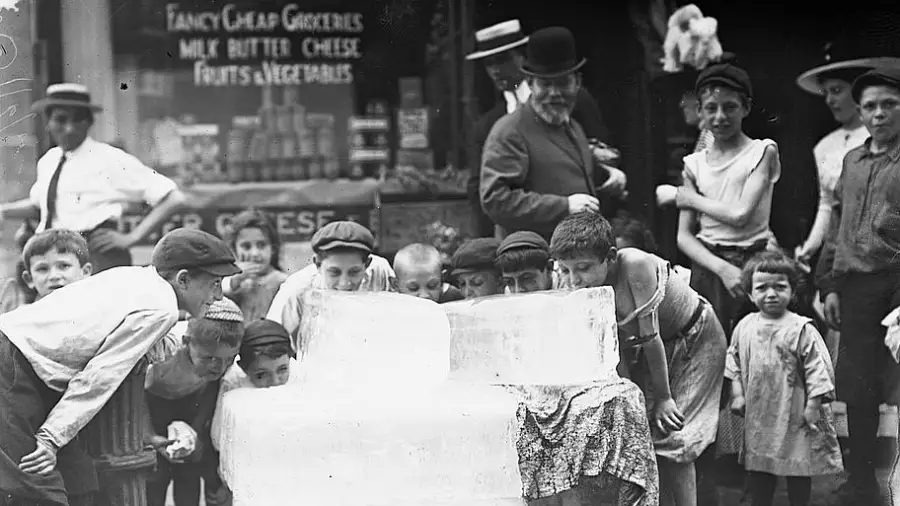 A group of children and a few adults gather around large blocks of ice on a street, touching and leaning close to the ice, with a grocery store sign visible in the background. The scene appears to be from the early 1900s.
