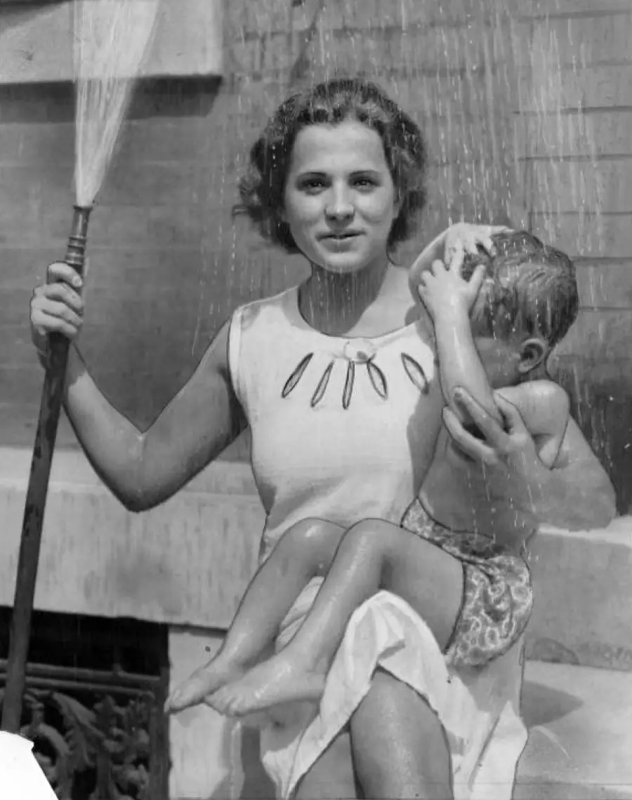 A woman in a sleeveless dress holds a broom in one hand and a small child in her lap while they sit outside under a spray of water, possibly cooling off on a hot day. The child covers their face.