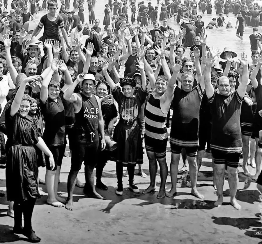A large group of people in old-fashioned swimsuits stand on a crowded beach, smiling and waving at the camera. Many wear dark, knee-length swimwear, and the beach is filled with people in the background.