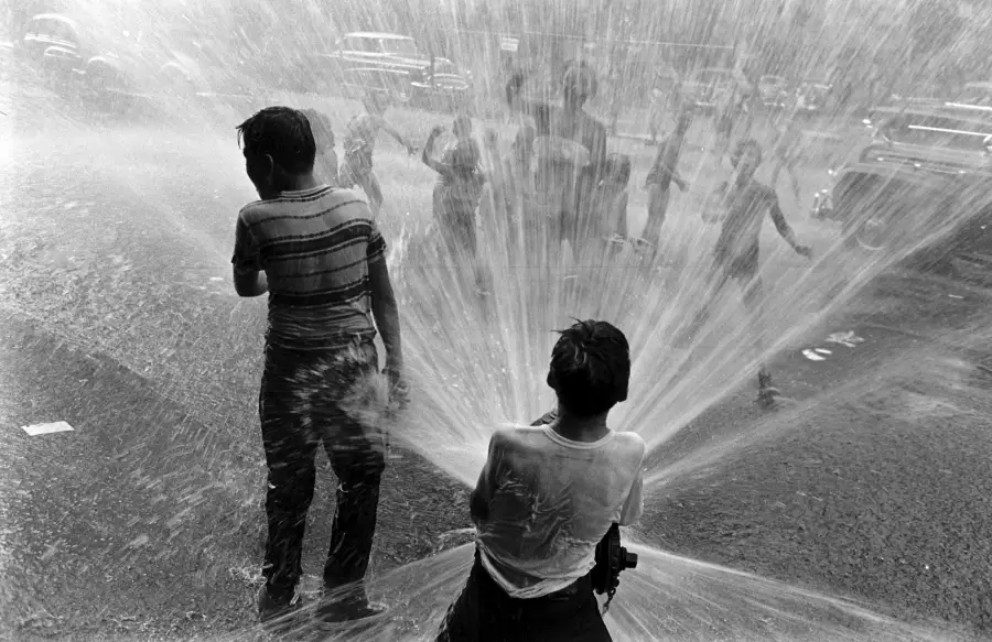 Children playing in the street, being sprayed by water from an open fire hydrant on a hot day, with cars parked in the background and water spraying everywhere.