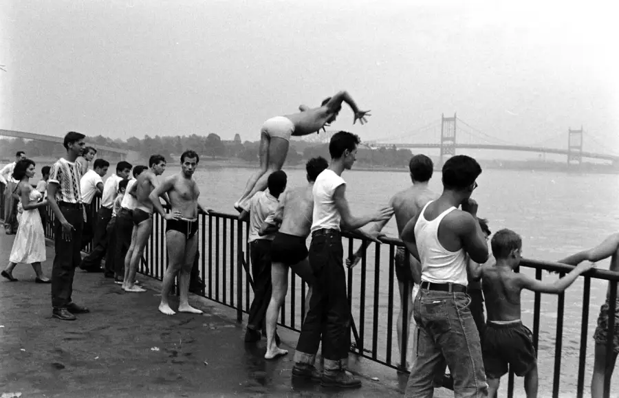 A group of people, mostly boys and young men in swimsuits and casual clothes, stand by a riverside railing while one boy dives into the water. A bridge is visible in the background on a cloudy day.