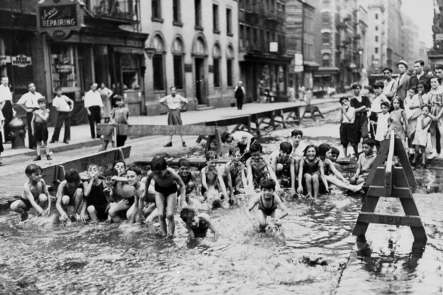 A group of children in swimsuits play and splash in water on a city street, surrounded by wooden barricades, while adults and other children watch from the sidewalks and buildings in the background.