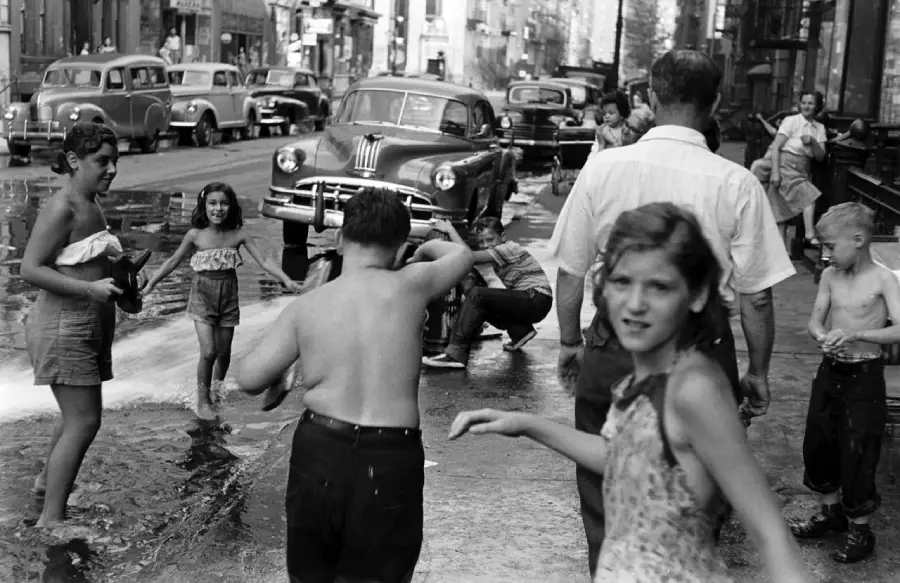 Children and adults play in water from an open fire hydrant on a busy city street lined with vintage cars and buildings, capturing a lively summer scene in an urban neighborhood.