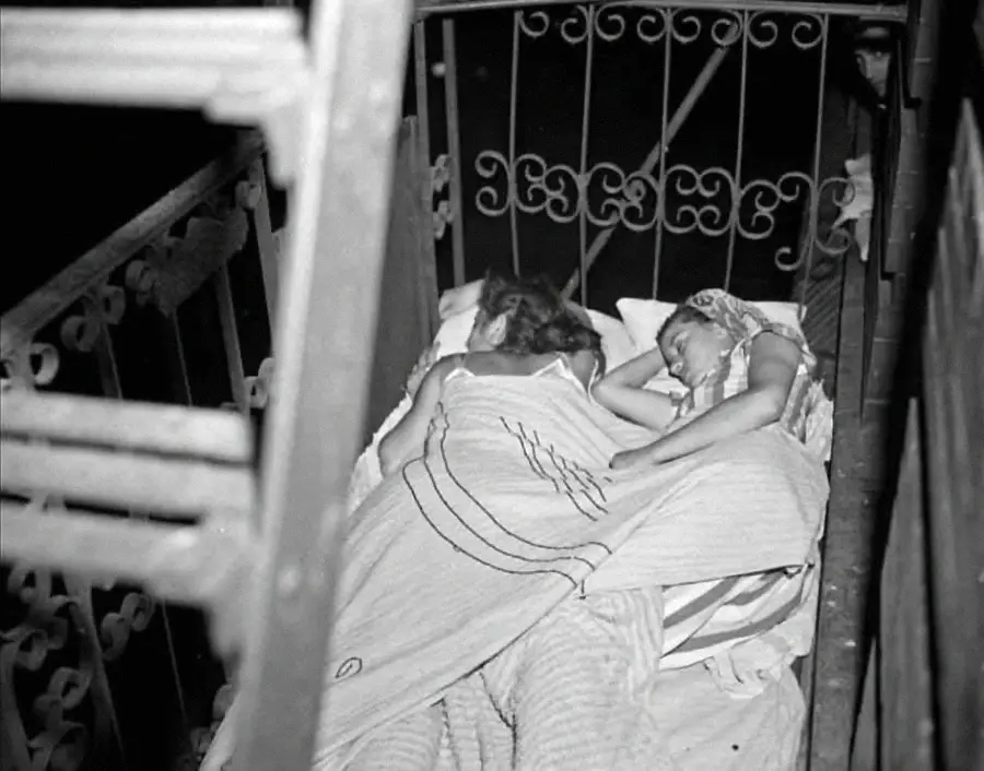 Two children sleep close together on a mattress outside, covered by a blanket, with wrought iron railings surrounding them and part of a staircase visible in the foreground.
