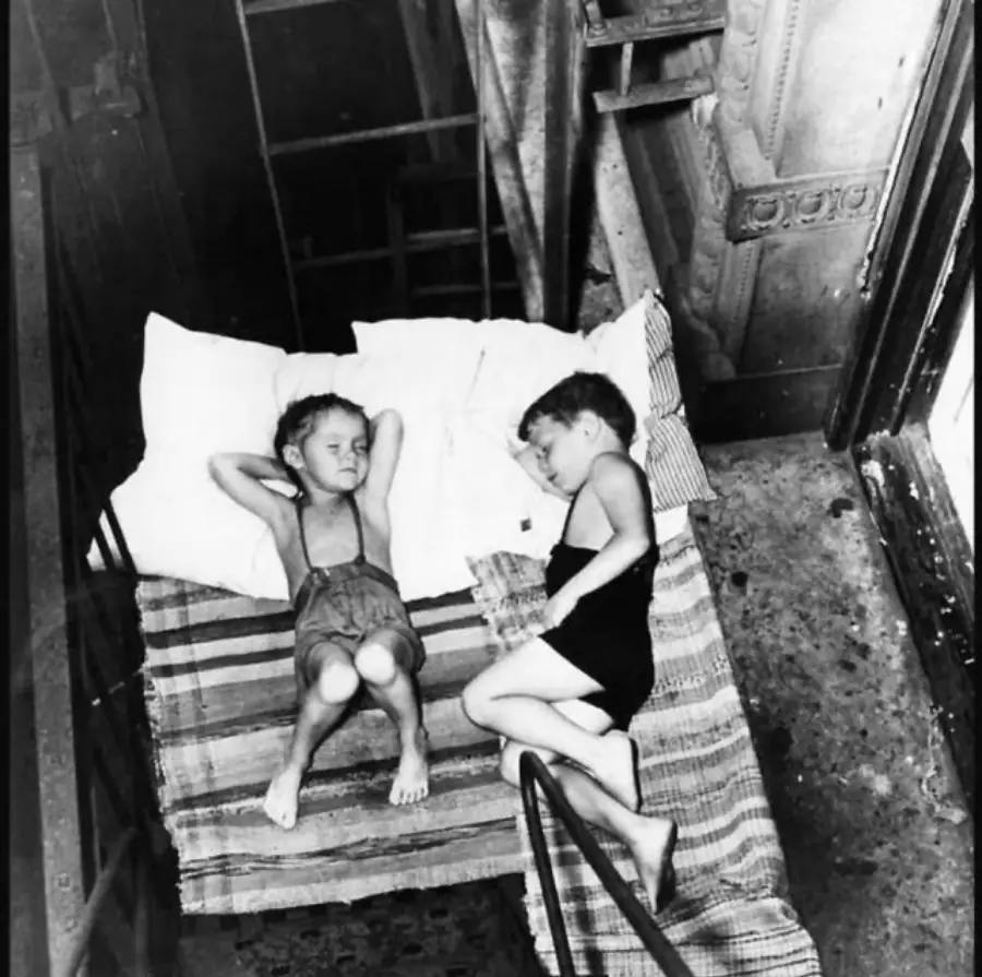 Two young children are sleeping on a mattress laid out on a fire escape. They lie next to each other with pillows and blankets, surrounded by railings and the exterior of a building. The scene is in black and white.