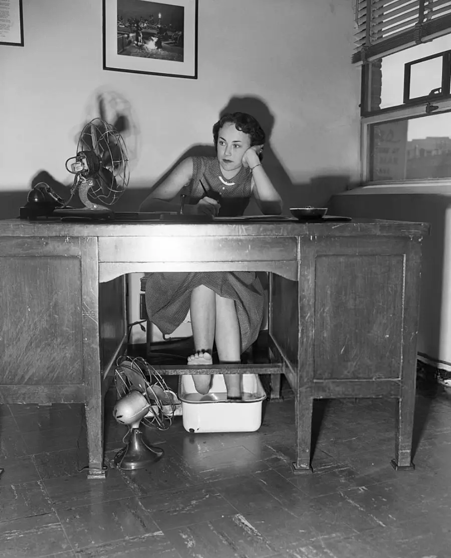 A woman sits at a desk, looking tired, with her feet in a basin of water and a fan blowing on her. Another fan is on the floor beneath the desk. Sunlight streams through a window behind her.