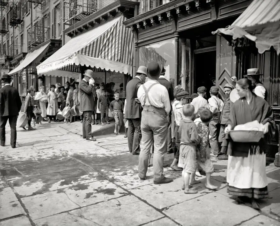 Black-and-white photo of people, including children, standing in line on a city sidewalk outside a storefront with striped awnings, likely early 20th century. The scene appears busy and lively.