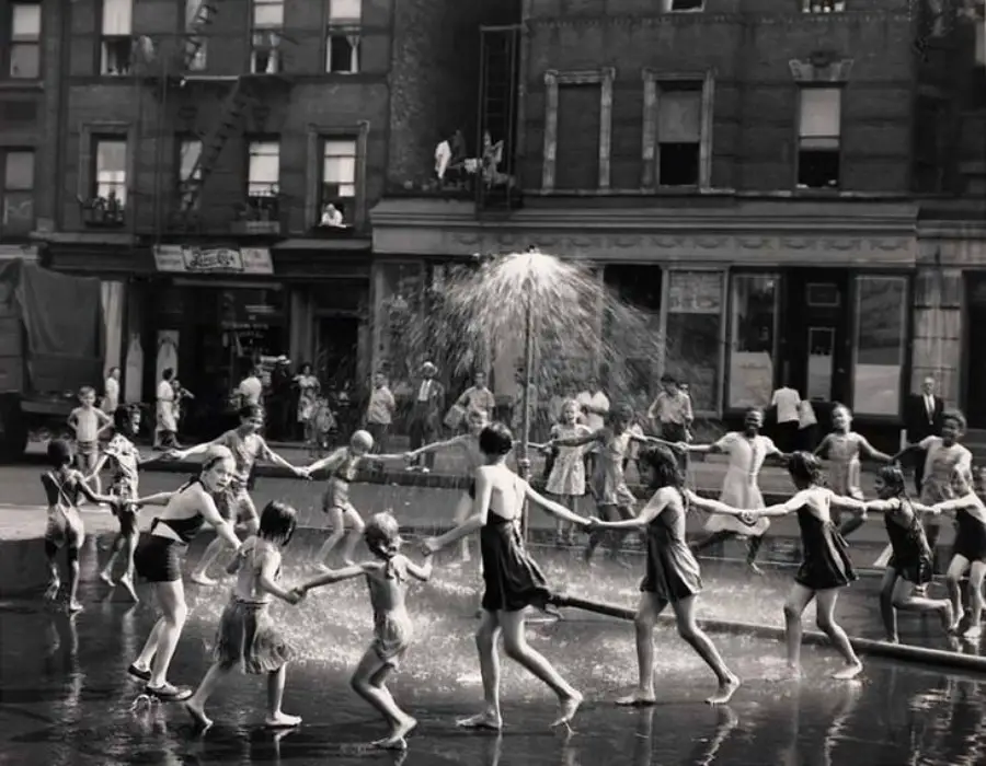 Children in swimsuits hold hands and play in a circle under a spraying street fountain, with apartment buildings and onlookers in the background on a sunny day.