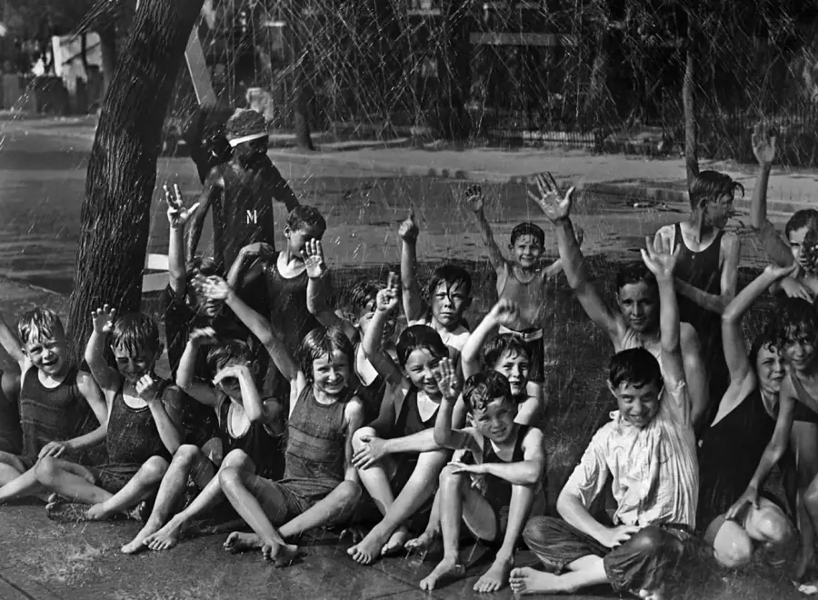 A group of children in swimsuits sits on a sidewalk, smiling and waving as water sprays down on them from above, creating a playful and joyful summer scene.