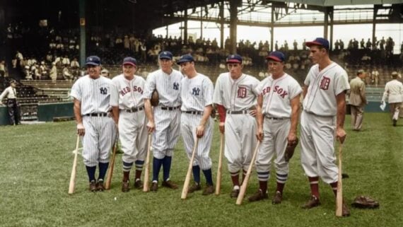Seven baseball players in vintage uniforms from the Red Sox, Yankees, and Tigers stand in a row on a baseball field, each holding a bat, with a stadium and spectators in the background.
