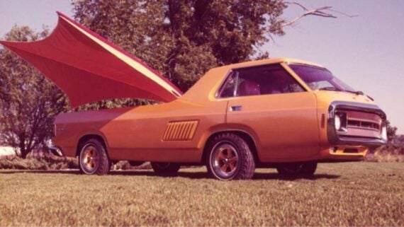 An orange vintage car with a unique design has a large, wing-like structure extending from its rear, parked on grass with trees in the background under a clear sky.