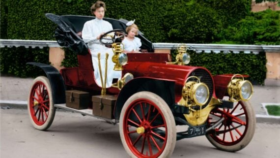 A woman and a young girl in white dresses sit in an old-fashioned red and black vintage car with gold details and large spoked wheels, parked outdoors in front of a green hedge.