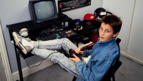 A boy in a denim jacket sits at a desk with his feet up, playing a retro video game on a small CRT monitor. The desk has caps, a helmet, and various electronic devices on it. The boy looks toward the camera.