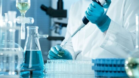 A scientist in blue gloves and a lab coat uses a pipette to transfer liquid into a small vial. Several other lab containers and equipment are visible in the background.