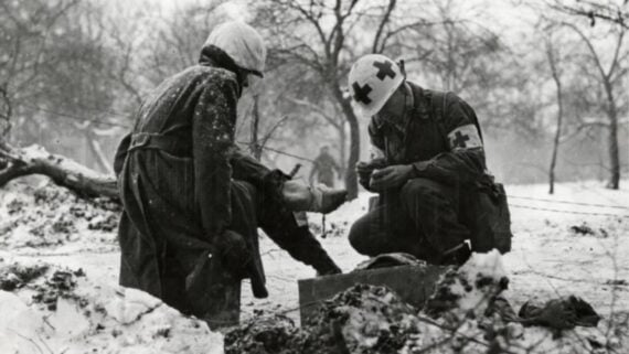 A medic wearing a helmet with red cross symbols bandages the leg of a wounded soldier in a snowy, winter battlefield with bare trees in the background.