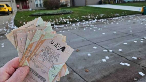 A hand holds several lottery tickets in the foreground, while numerous paper slips are scattered across a residential driveway and front lawn in the background.
