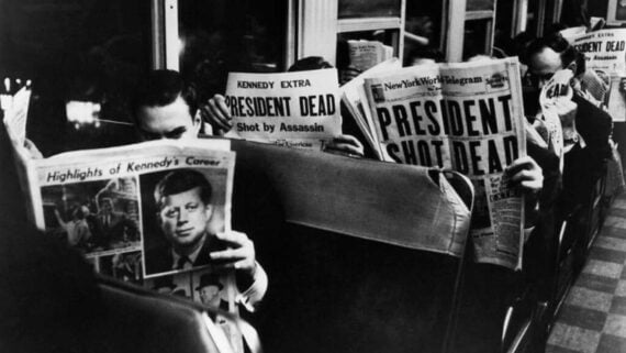 Several men sit on a bus reading newspapers with bold headlines announcing President Kennedy&rsquo;s assassination. The atmosphere appears somber as the shocking news is displayed prominently on the papers.