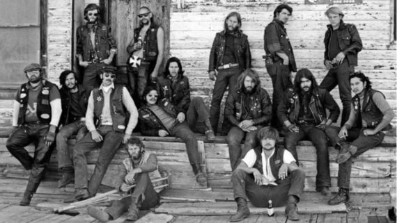 A group of men wearing leather vests and jackets pose together outside a weathered wooden building. Some stand while others sit on the porch steps or the ground, looking directly at the camera.